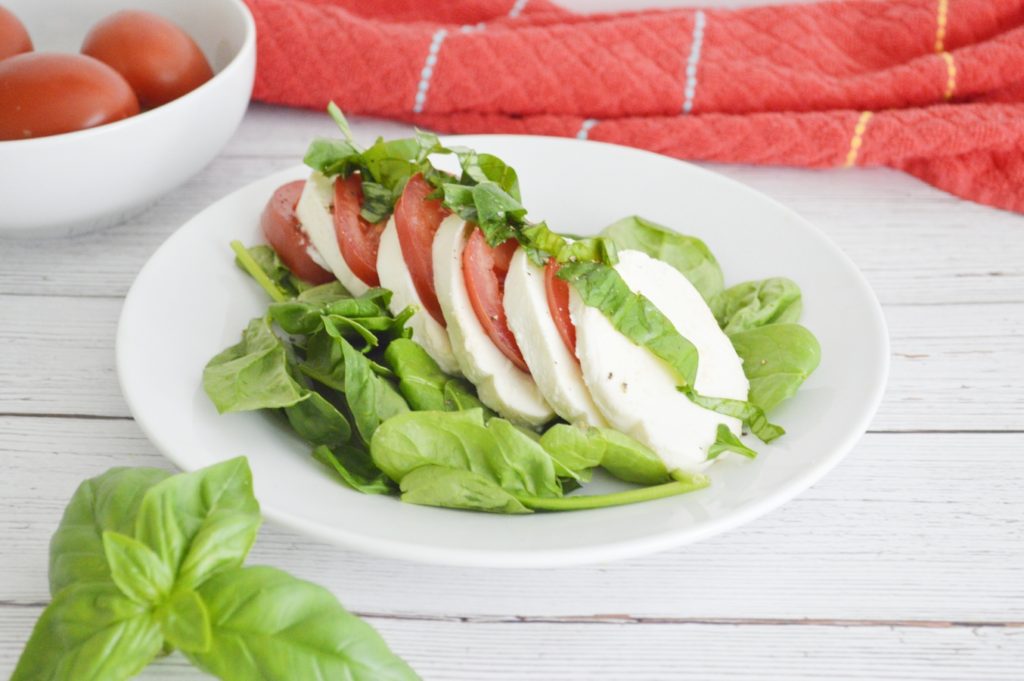 Easy Caprese Spinach Salad on white plate with basil and tomatoes in background
